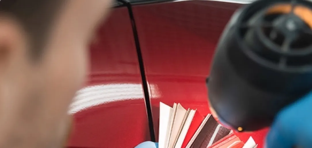 A technician holding paint color swatches against a red car panel under a work light
