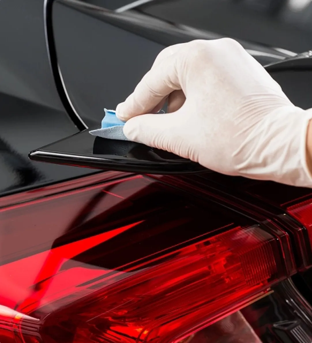 A gloved hand polishing the edge of a black car panel above a red tail light